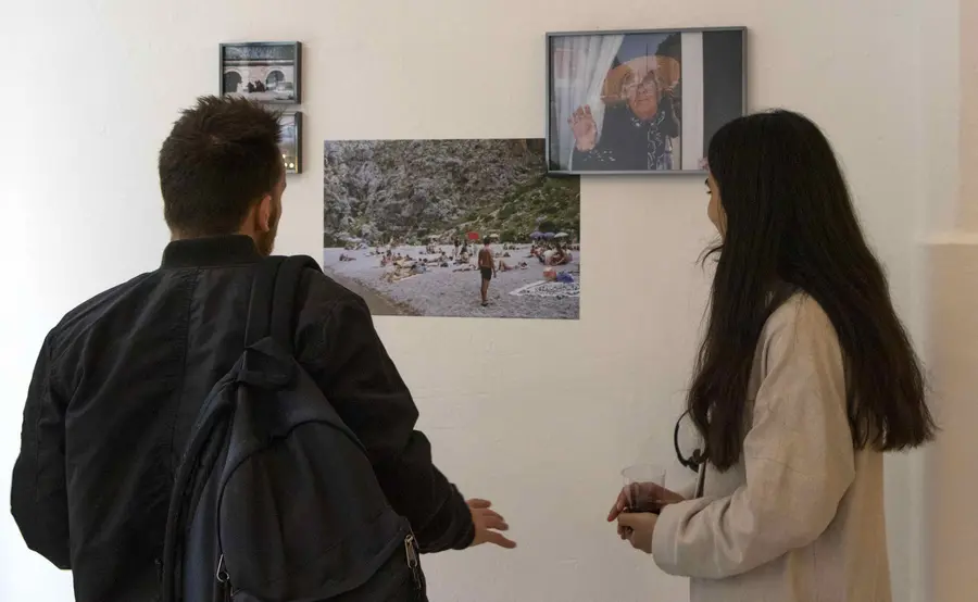 A photograph of 2 people look at art work displayed on a white wall.