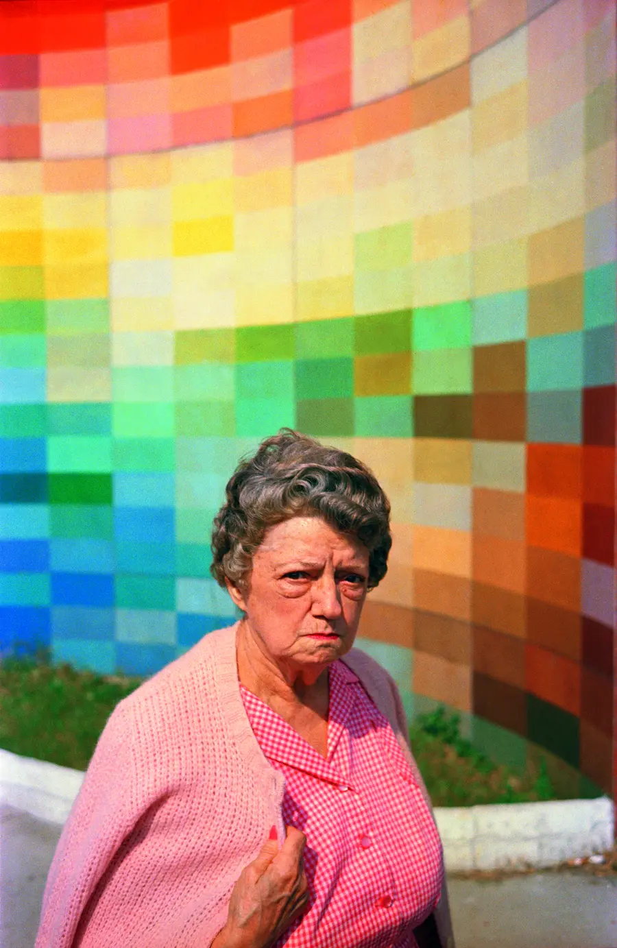 Colour photograph of an elderly woman wearing a pink blouse and cardigan standing in front of a multi-coloured background.