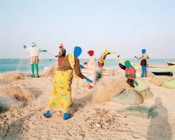 A group of people on a sandy shore are at work untangling fishing nets. They are dressed in shorts or saris, their heads and limbs are painted solid colours of either yellow, red, blue or green.