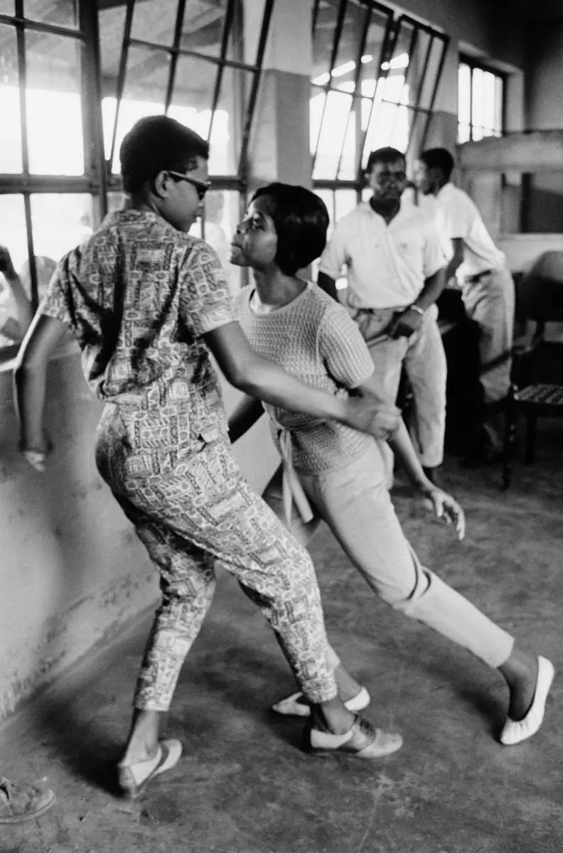 Black and white photograph a man and woman dancing together in a room. Two other men are stood in the distance.