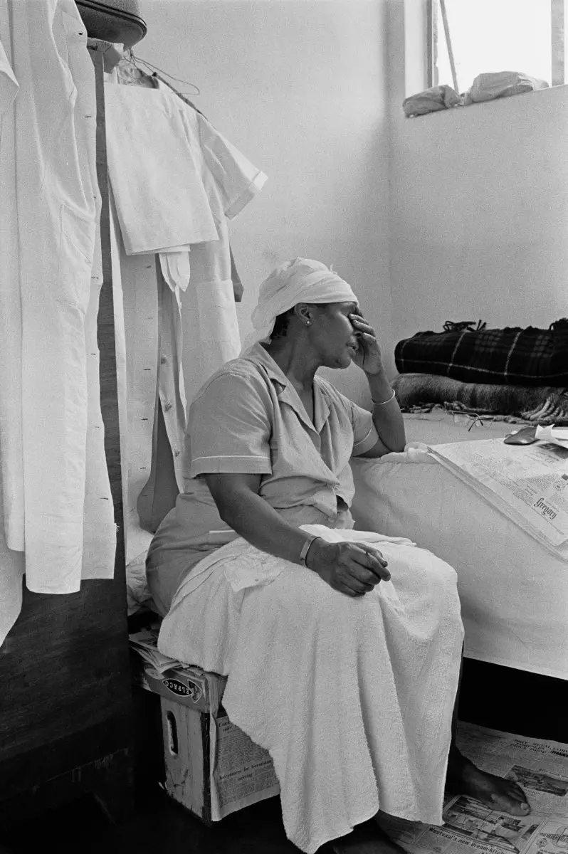 Black and white photograph of a black woman in cleaning scrubs sat on a crate as she leans her head in her hand. A white cloth is strewn over her lap.