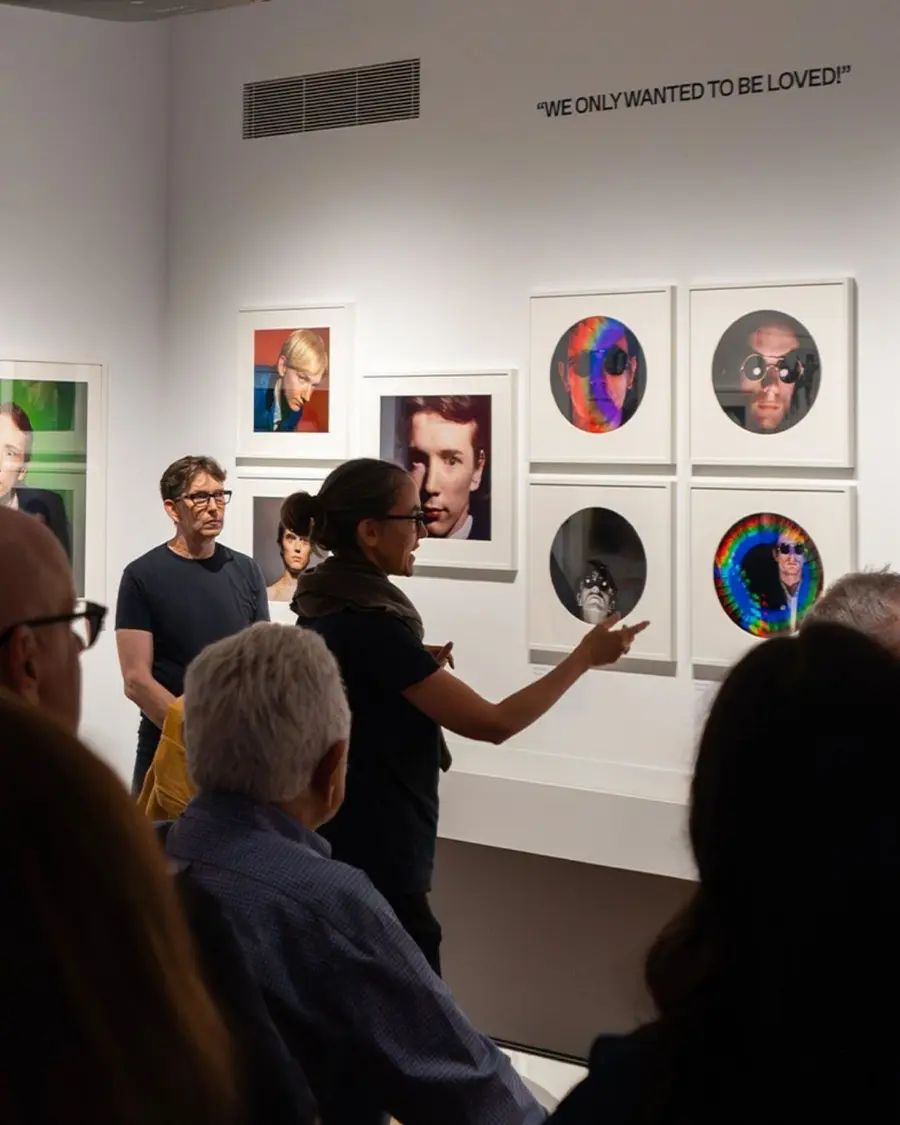 A curator leads a tour in an exhibition space. She raises her hand to indicate to a displayed photograph while a tour group looks on with interest.