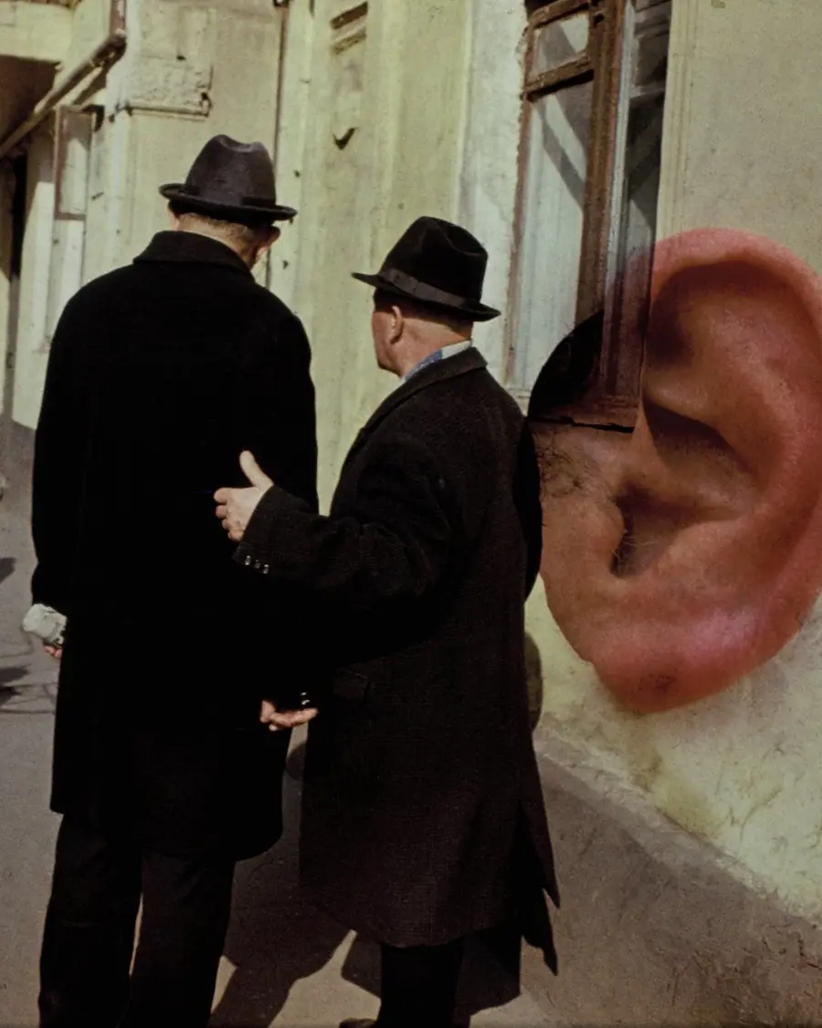 Colour photograph of the backs of two men  standing in the middle of a pavement. Layered on top is the  image of an ear