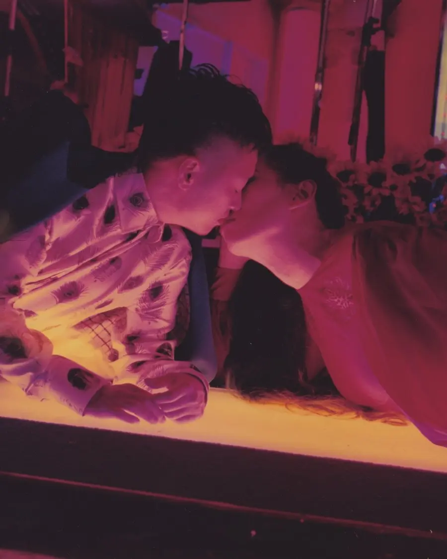 A darkened bar lit with a pink glow. A couple lean across it to meet in a kiss.