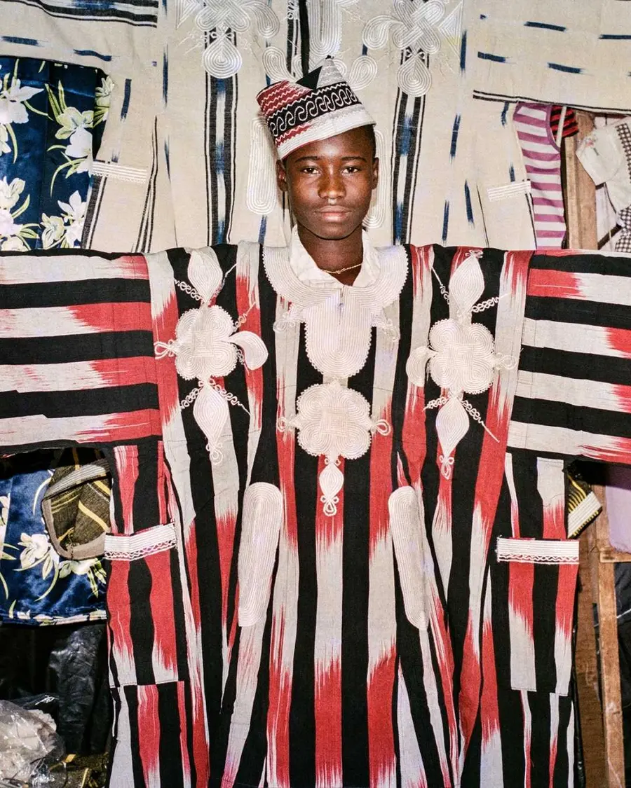 A young man dressed in a black, white and red striped traditional tunic poses with his arms spread wide open in a studio.