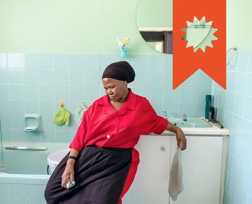 Colour photograph of a woman sat in a bathroom looking down. She holds a sponge in her hand.