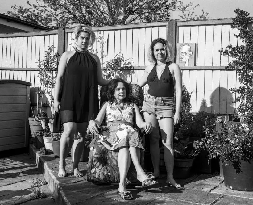 A black and white photo of three women in the same family of different generations sat for a portrait in a driveway. The youngest sits on an outdoor garden chair whilst the two elder women stand behind her by each shoulder. They look content and relaxed.