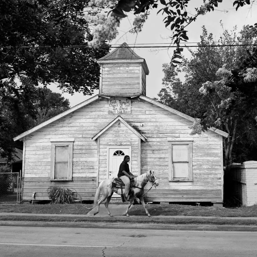 Black and white photograph of a man riding a horse past a house on the street.