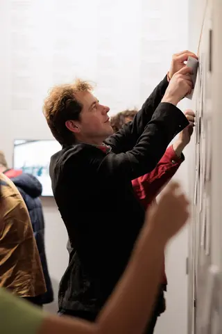 Colour photo of a man in a dark blazer pinning a page to a wall in an exhibition space.