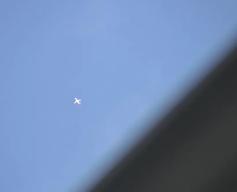 A small plane flights against the blue sky, the edge of a what looks like a blurred building covers the lower right corner of the image