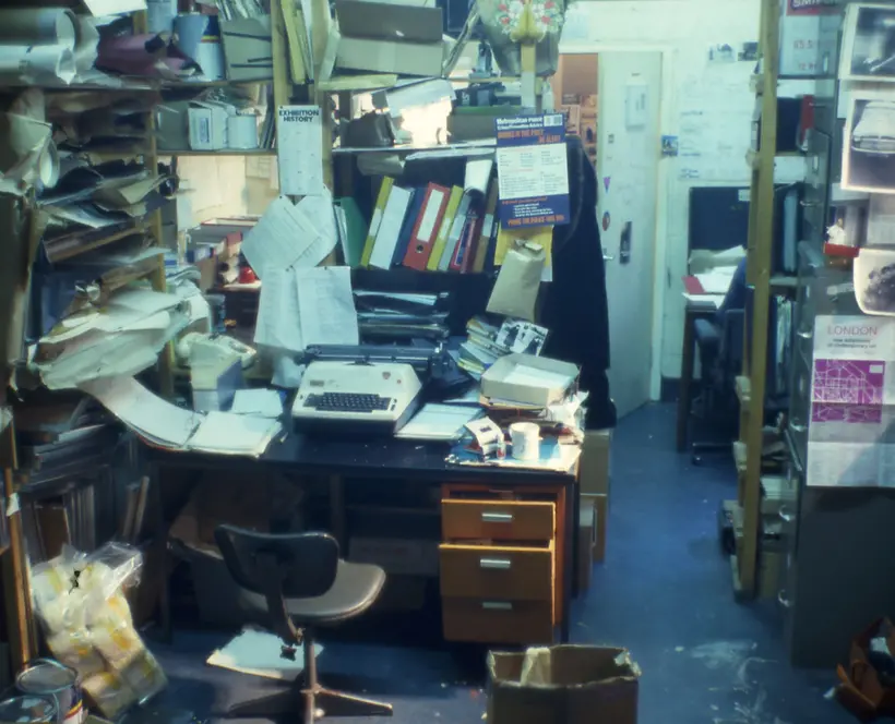Archival full colour photograph with a desk in the middle of an office surrounded by papers, books, binders and a miscellany of other office supplies and materials