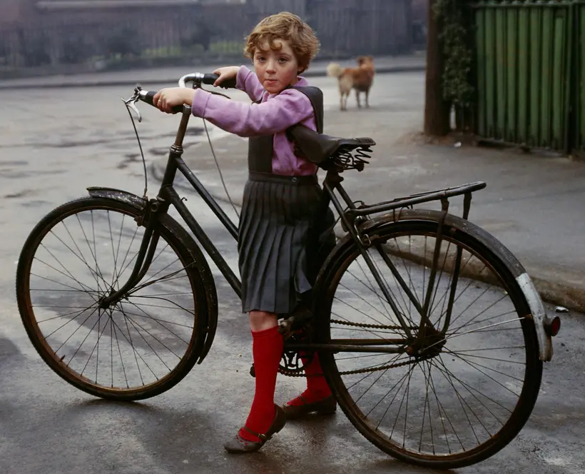 Colour photograph of a small child, straddling the frame of an adult-sized bicycle, who has stopped on a street and turned to look at the photographer.