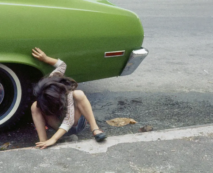 A child crouches on the pavement next to the back tyre of a green car