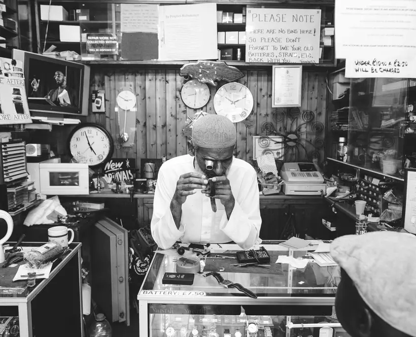 Black and white image featuring a man in a shop leaning on a vitrine surrounded by clocks