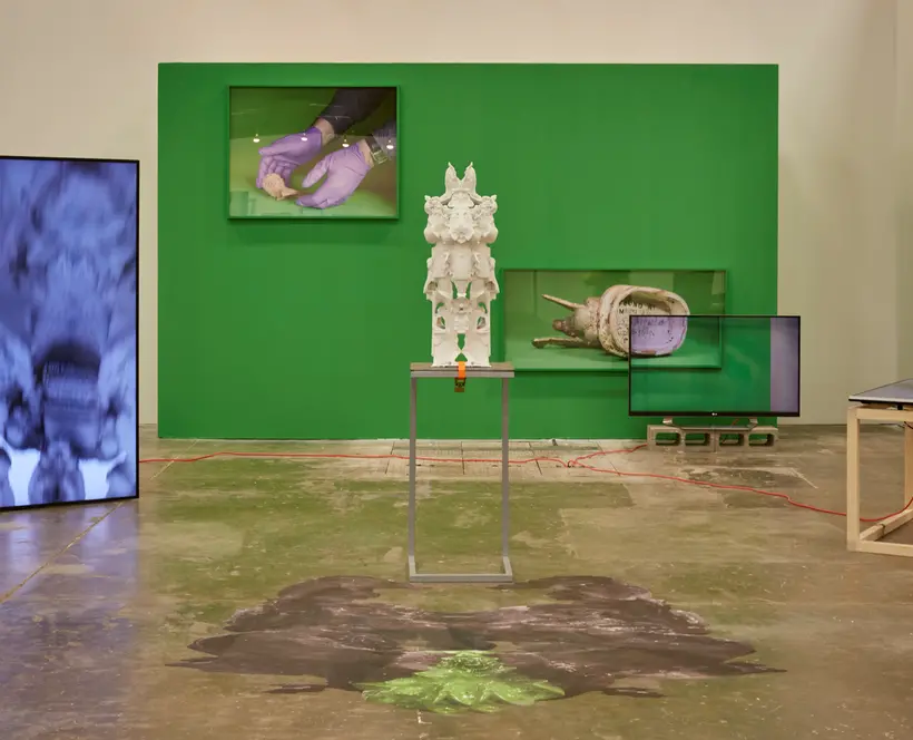 An installation view of an exhibition on a gallery. There's a sculpture in the middle of the image, a built wall painted in green in the back, a large vertical screen on the left and other art works on view.