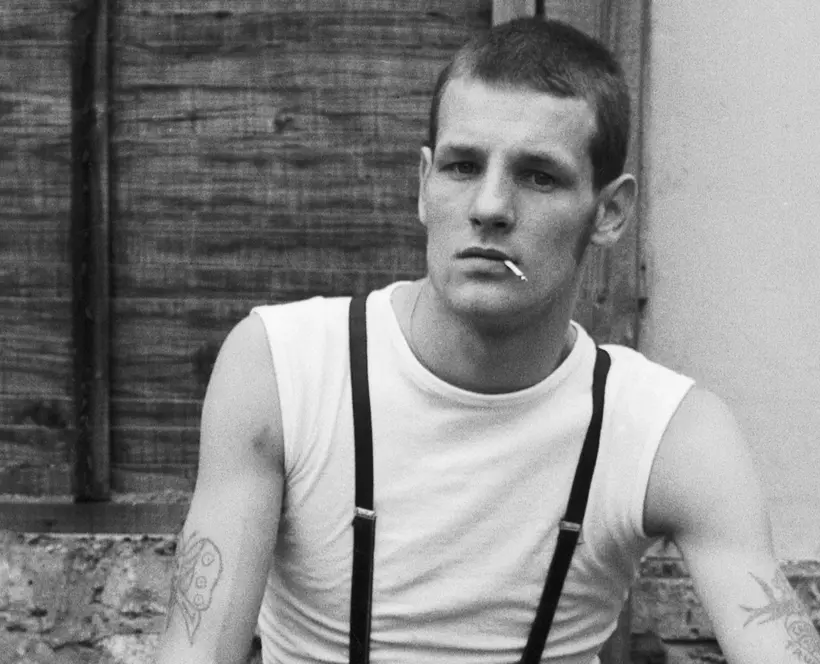 Black and white photograph of a young man in a white sleeveless vest and braces, short cropped hair and cigarette dangling from mouth, staring into at the camera.