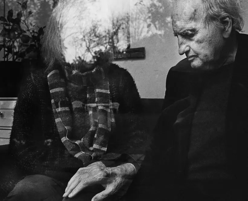 Black and white image featuring an older couple with the person on the right's face partially obscured by a reflection on glass. Man's hand is on the other person's knee.