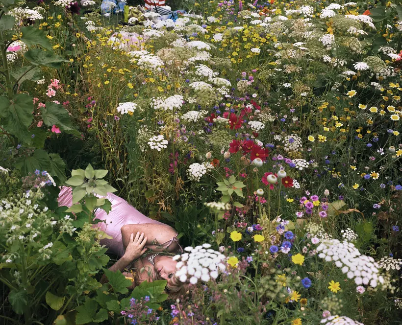 Colour photograph of a woman lying down with her arms wrapped around herself in a garden full of flowers.