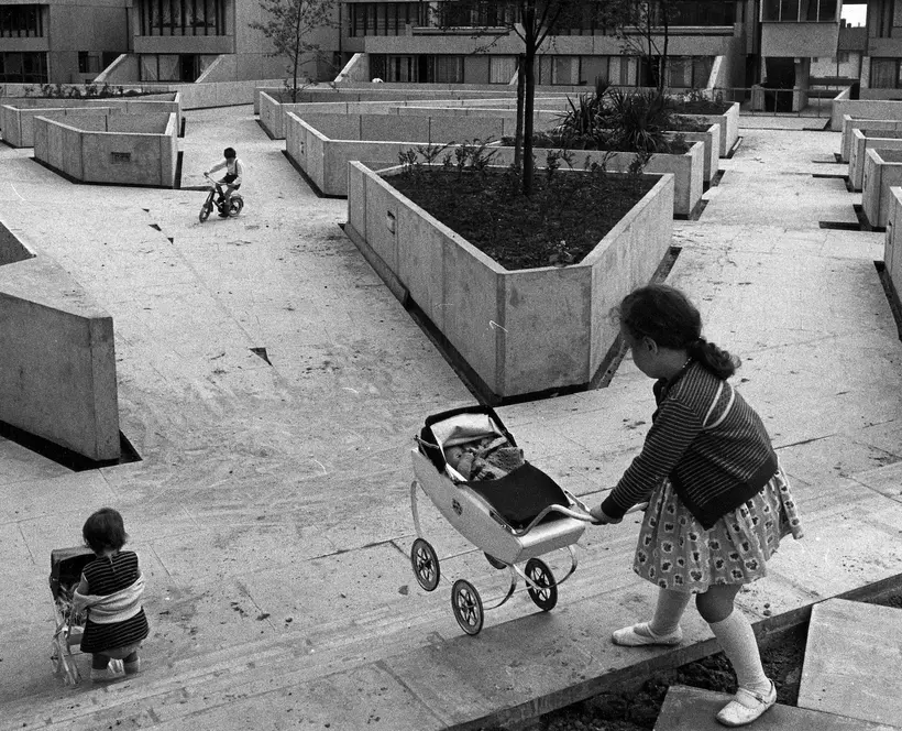 A black and white photo showing two children pushing prams on stairs in a concrete courtyard while another cycles in the distance.