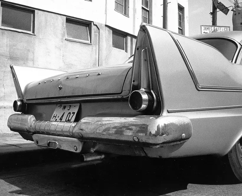 Black and white photograph of the rear of a 1960s car on a street with a person walking past