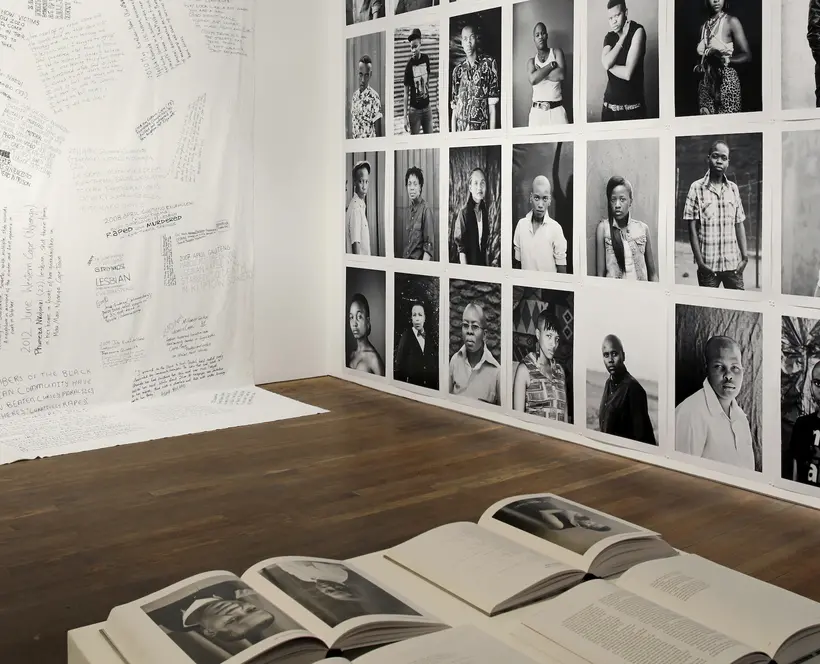 The corner of an exhibition space with a photography book on a plinth in the foreground, black and white portrait photographs along one wall and handwriting on a white background along the other wall. 