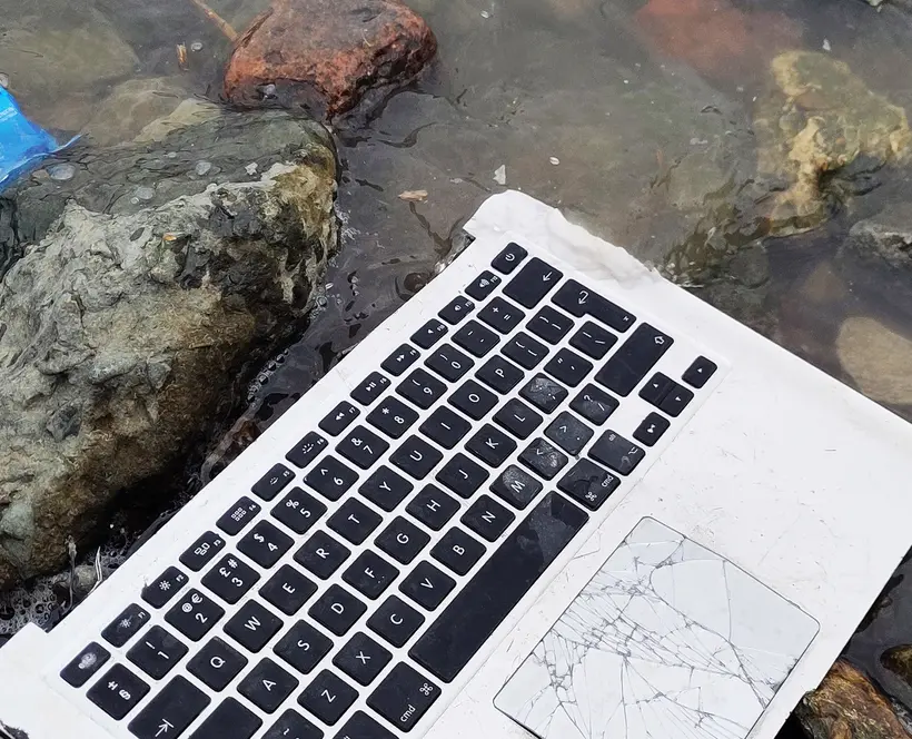 A broken laptop keyboard and an empty bag of crisps sitting ontop of a rocky foreshore.