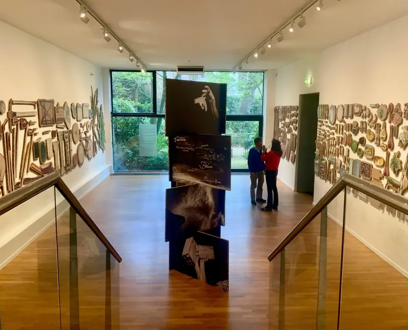 An exhibtion room with a large glass window on the far side. Photographs are stacked in a column in the middle of the room, objects printed on wood hang on the wall 