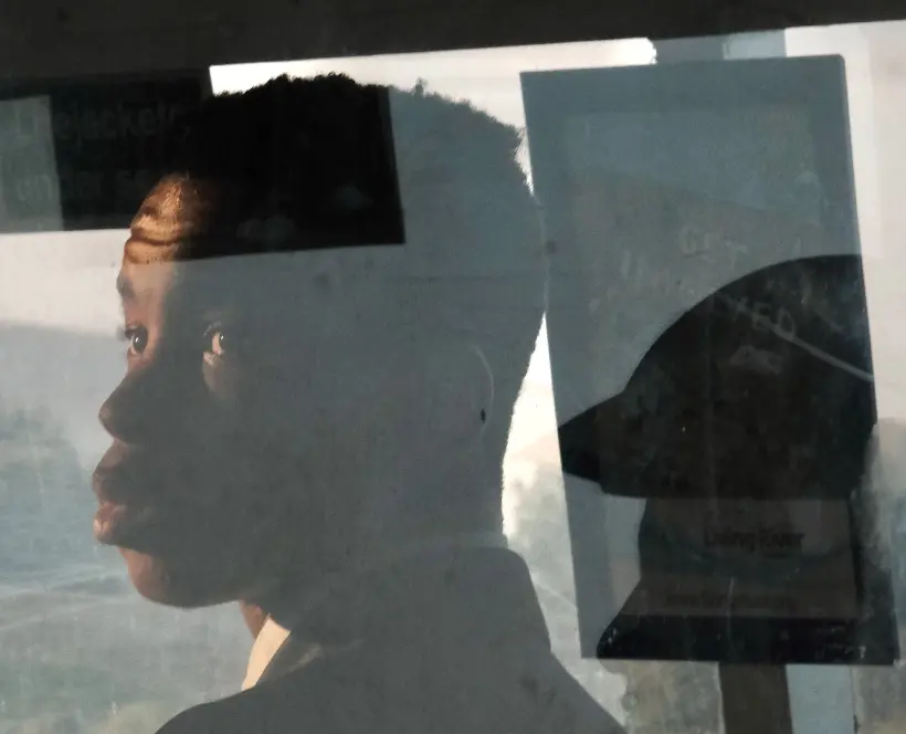 Colour photograph of a young person sat behind a clear partition, looking over their shoulder towards the camera. In the glass there is a reflection of another person