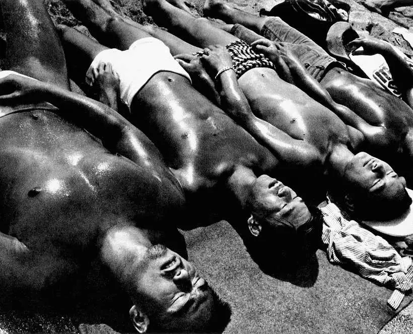 Black and white photograph of a group of men sunbathing, laying in a row next to each other wearing swimming trunks