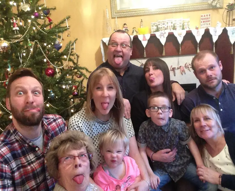 An image of three generations of a family, all pulling silly faces for the camera. A Christmas tree is in the background.