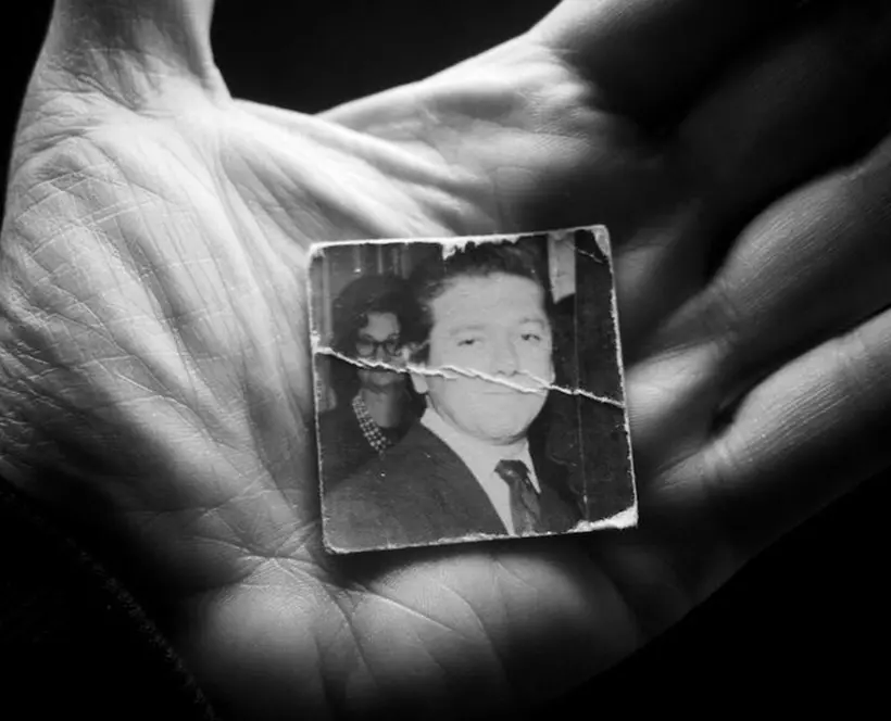 A hand holds a small square photo of a person's father