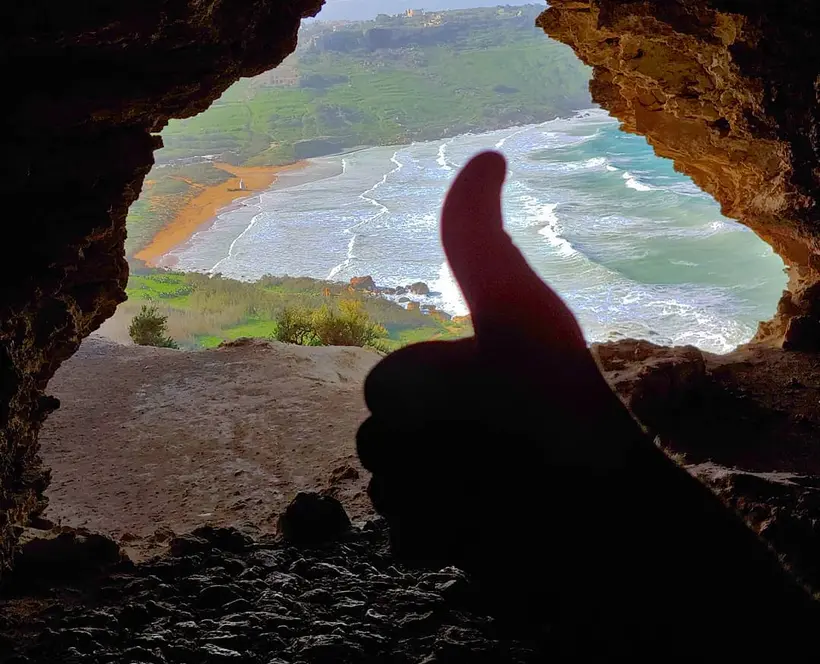 A photograph of a beach taken from the entrance of a cave. A hand with the thumb up is in the foreground.