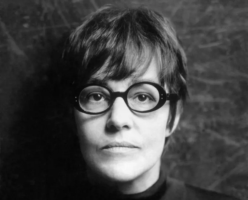 Black and white photograph of a woman looking directly at the camera, her head and shoulders can be seen. She is wearing black rimmed glasses, she has short, dark hair and is wearing a black jumper.