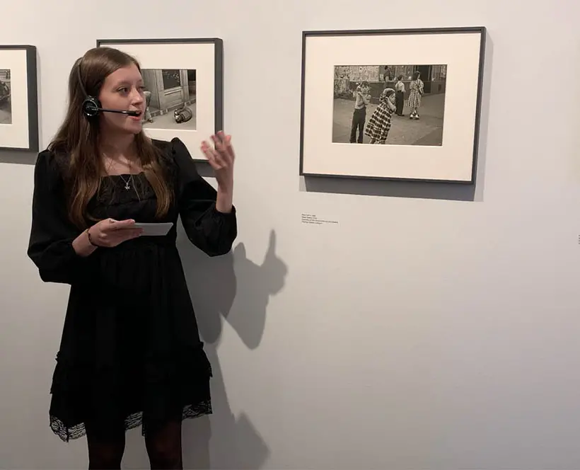 A person standing in the left of the frame presenting in front of photographs in frames on walls.