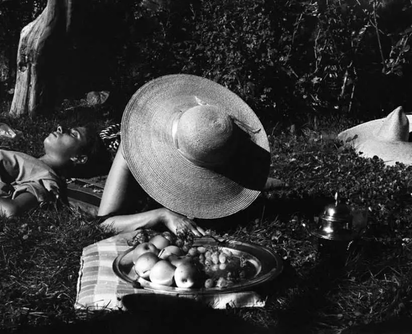 Black and white photograph of people laying on the grass with a picnic. Person in the centre of the image has their head lowered, showing a straw sun hat