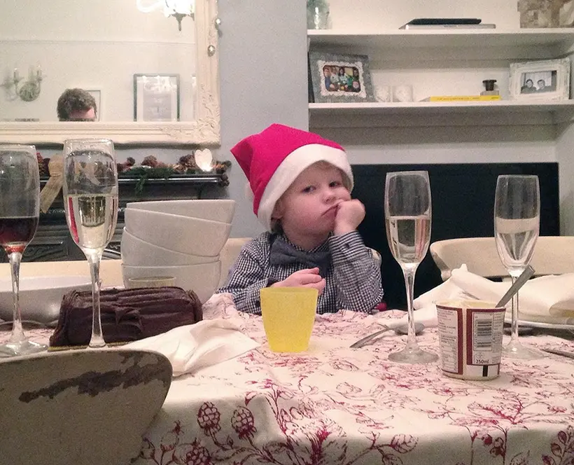 A photograph of a small child with a Christmas hat on, sat at a Christmas dinner table looking tired and a bit grumpy, head in his hand.