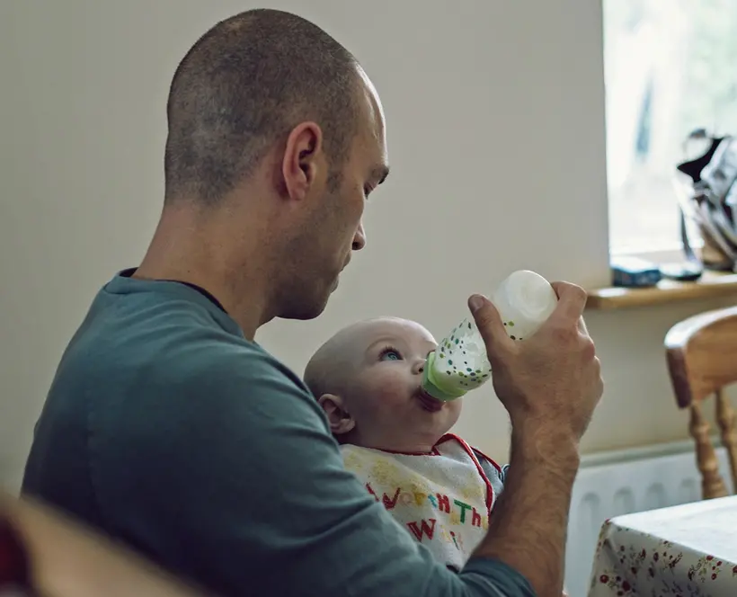  A picture of a father bottle-feeding his baby while sitting at a kitchen table