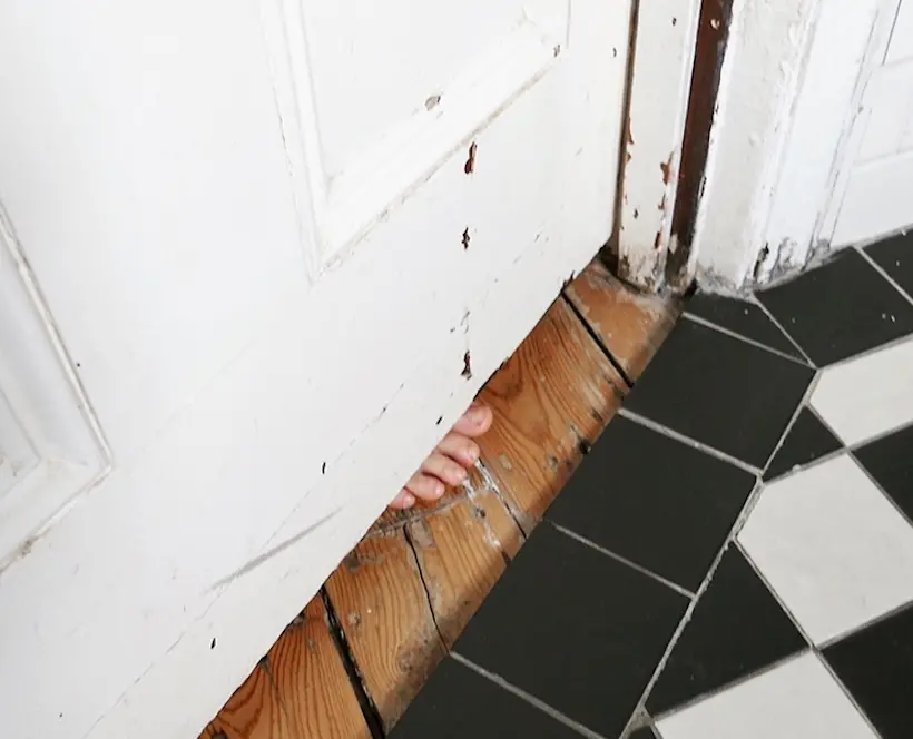 A photograph of a closed bathroom door with a child's toes poking underneath it