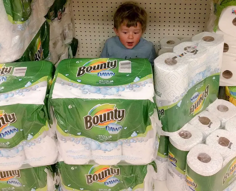 A photograph of a little boy's face peeking out from a mountain of toilet roll in a supermarket