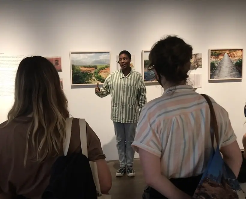 A photograph of person in the centre speaking to a crowd in a gallery.