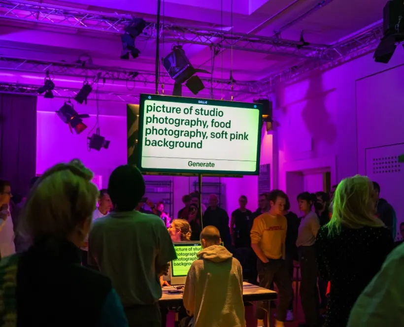 A pink lighted crowded space with a big screen high on the middle of it. On the screen it says" Dall-E. Picture of studio photography, food photography, soft pink background. Generate". Under the screen a person writes types those letters on a computer.